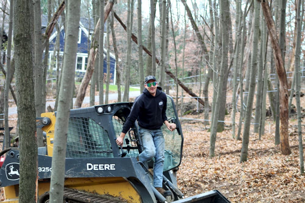 Man operating a John Deere skid steer in a wooded area with autumn leaves.