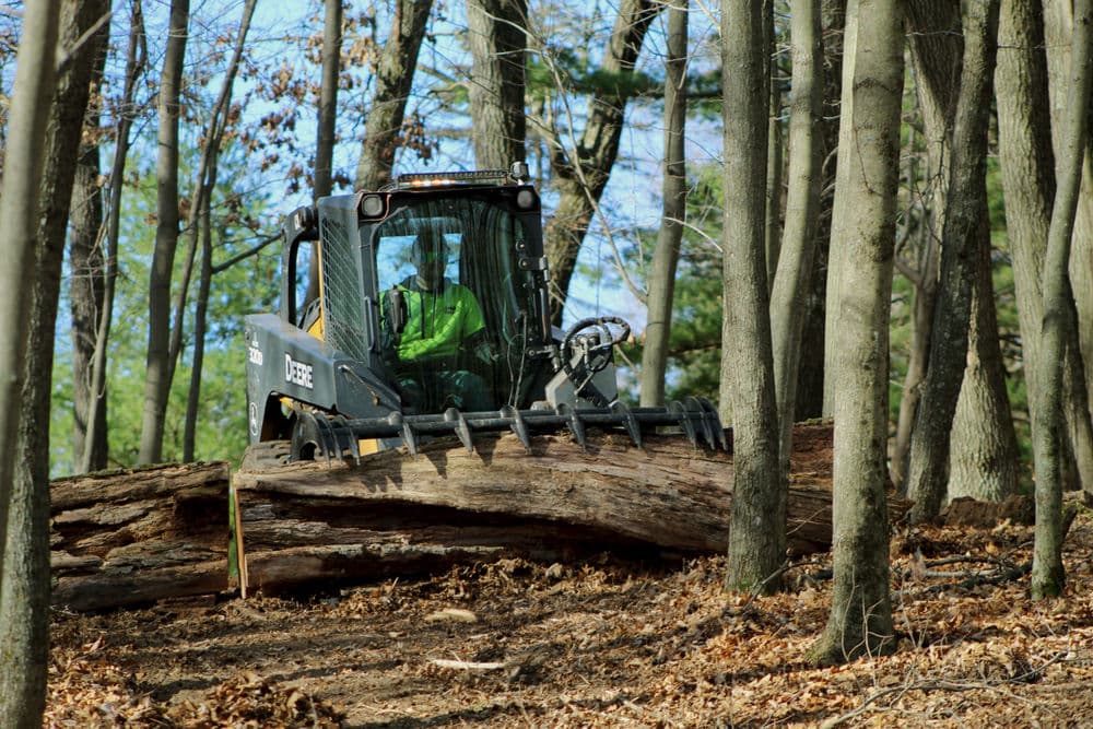 Skid steer loader moving a large log in a wooded area with worker visible inside.